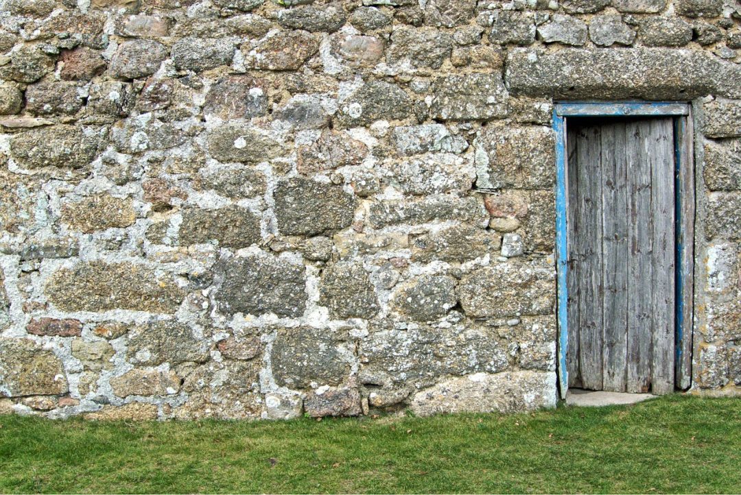 The image depicts a weathered gray wooden door set into a buildings hand laid stone wall. 