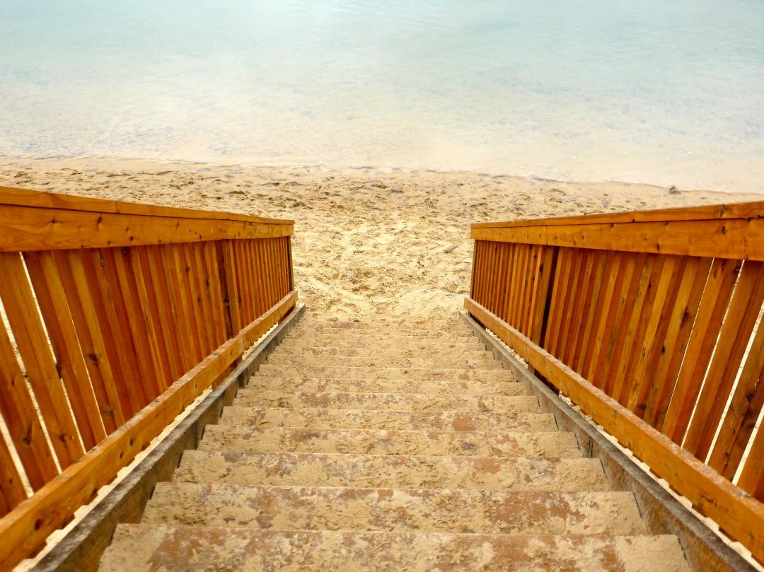 The image depicts a steep wooden staircase with many steps descending to a beautiful sandy beach with the water beyond the beach.